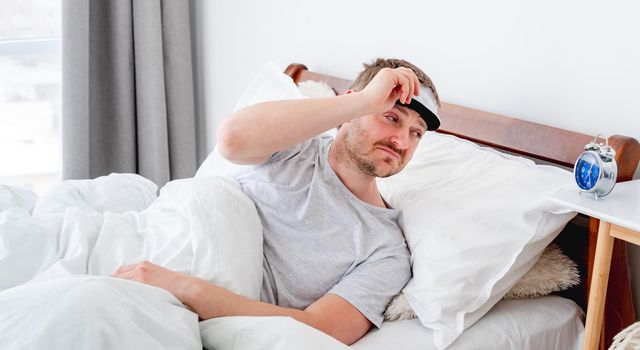 Man wearing pajamas and eye sleeping mask waking up in the bed in the morning time and looking at the clock. Guy resting in the room with daylight. Male person under blanket at home