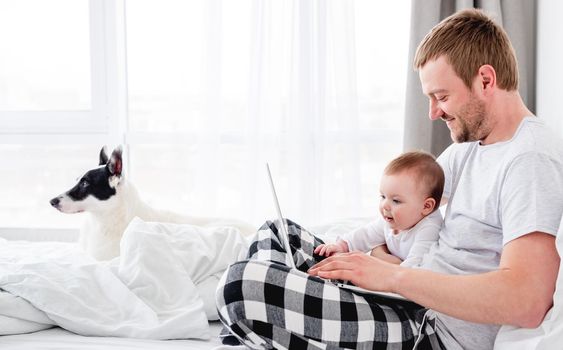 Young father sitting in the bed with little son and cute dog and working on notebook. Man freelancer staying in the bedroom with his child boy and pet doggy in the morning time