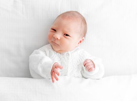 Adorable newborn baby girl wearing white costume lying in the bed at home with daylight and looking back. Portrait of cute infant child under blanket