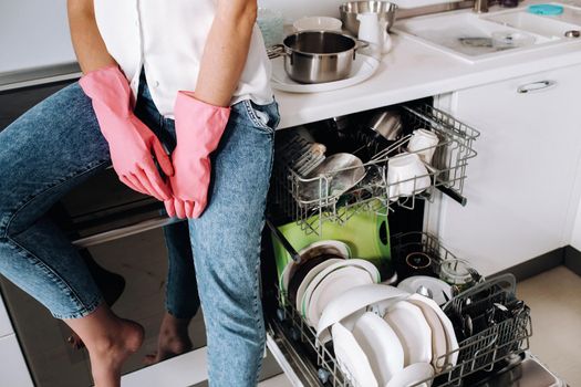 a housewife girl in pink gloves after cleaning the house sits tired in the kitchen.In the white kitchen, the girl has washed the dishes and is resting.Lots of washed dishes.