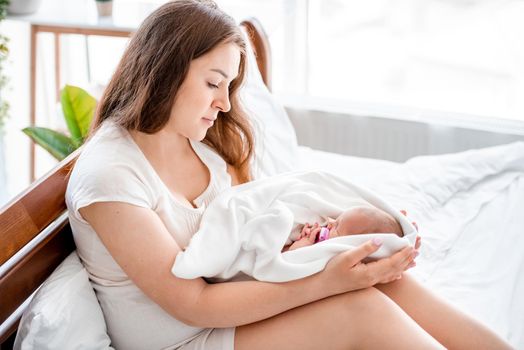 Beautiful young mother sitting in the bed and holding newborn baby daughter sleeping in white sheets. Pretty woman mom with her infant child girl at home