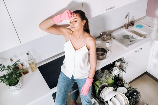 a housewife girl in pink gloves after cleaning the house sits tired in the kitchen.In the white kitchen, the girl has washed the dishes and is resting.Lots of washed dishes.