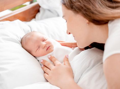 Young mother looking at her sleeping newborn daughter swaddled in white sheets at home. Portrait of girl mom with her infant child napping in the bed. Matherinity moments