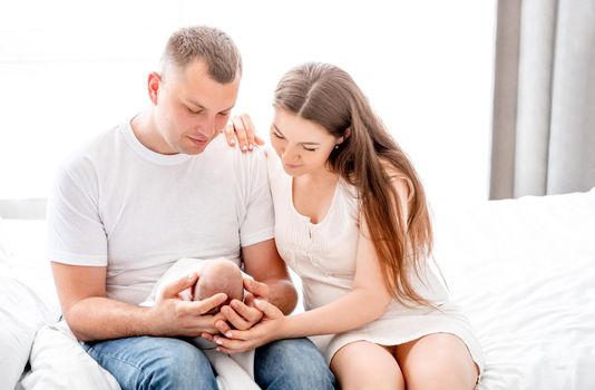 Beautiful young family sitting in the bed and holding newborn daughter in the room with daylight. Adorable infant baby girl sleeping in hands of her mother and father at home. Family portrait indoors