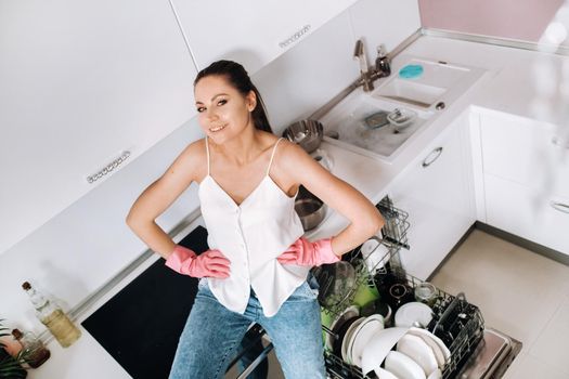 a housewife girl in pink gloves after cleaning the house sits tired in the kitchen.In the white kitchen, the girl has washed the dishes and is resting.Lots of washed dishes.