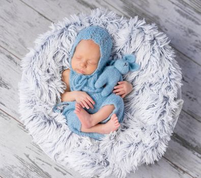 lovely infant in hat and jumpsuit sleeping on round little bed, top view