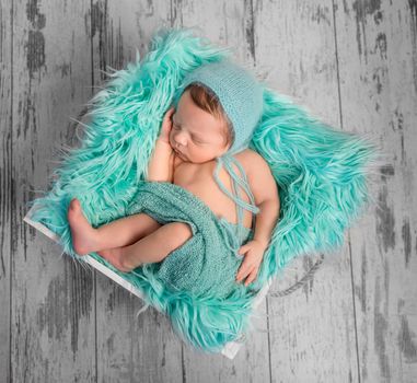 beautiful newborn baby sleeping in hat on square bed with furry turquoise blanket, top view