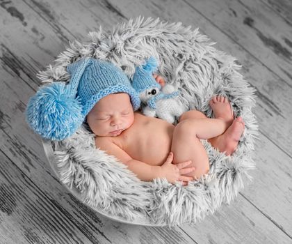 lovely newborn in blue hat with pompom and toy on round bed with furry blanket, top view