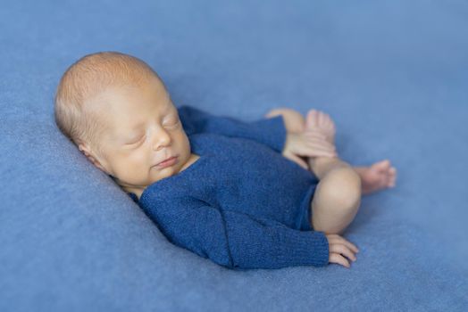 sleepy newborn baby in blue jumpsuit smiling and holding his leg