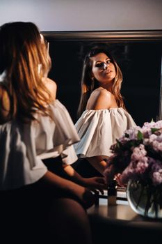 Beautiful girl in the mirror reflection at home. Girl before the holiday near the home mirror.A girl in a white dress with long hair is going to a party at home.