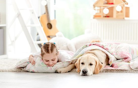 Happy little girl sleeping with lovely dog under blanket on floor in light room