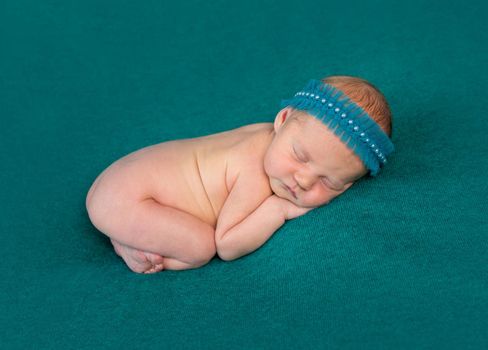 sweet naked newborn sleeping on stomach and hands with headband on dark turquoise blanket