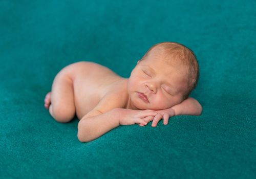 sweet naked newborn sleeping on stomach and hands with headband on dark turquoise blanket