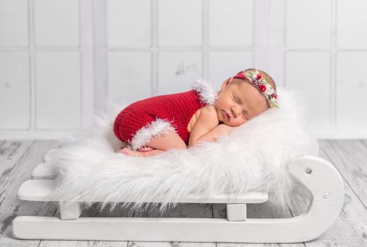 beautiful newborn in red romper sleeping on sleigh cot with white fluffy blanket