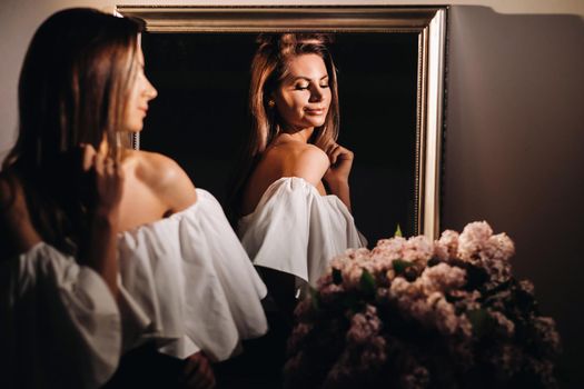 A beautiful girl in the mirror is reflected at home. Girl before the holiday near the home mirror.A girl in a white dress with long hair poses near a large mirror at home.
