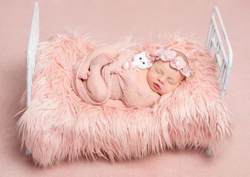 cute sleeping newborn girl with crossed legs and toy cat on little bed with pink fluffy blanket