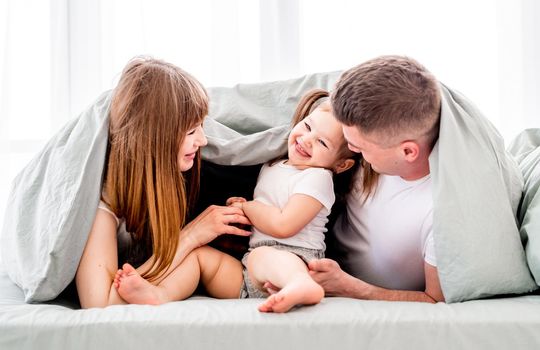 Family under the blanket in the bed. Young parents playing with small daughter under coverlet and smiling. Beautiful girl with her husband and kid in the bedroom