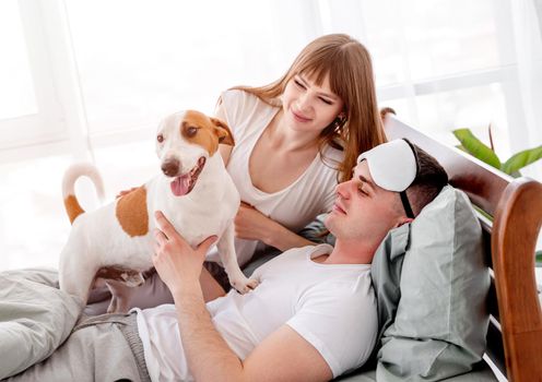 Beautiful couple lying in the bed with cute dog. Young woman and man wake up in the morning and petting their doggy. Caucasian family wearing pajamas in the bedroom with pet