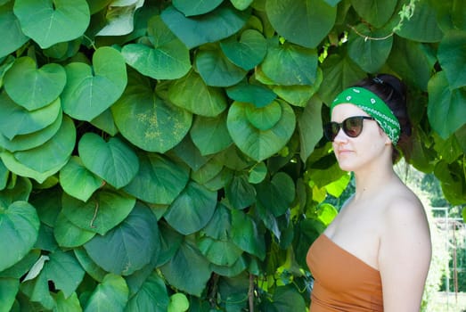 young woman on a background of a summer green park, green leaves. A girl with black hair, a green bandage on his head, in a brown tank top and black sunglasses.