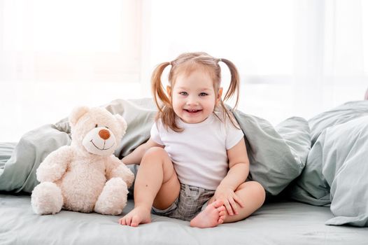 Little girl with teddy bear sitting in the bed and smiling. Beautiful portrait of cute child in the morning time in room with sunlight. Happy kid with toy