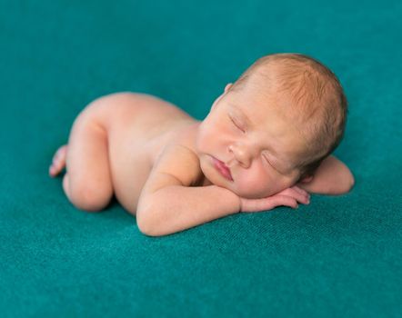 sweet naked newborn sleeping on stomach and hands with headband on dark turquoise blanket