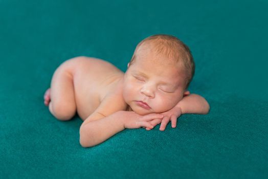 sweet naked newborn sleeping on stomach and hands with headband on dark turquoise blanket