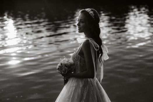 An elegant bride in a white dress and gloves stands by the river in the Park with a bouquet, enjoying nature at sunset.A model in a wedding dress and gloves in a nature Park.Belarus. black and white photo