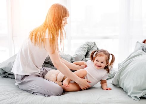 Beautiful mother playing with little daughter in the bed in the room with sunlight. Happy family moments of mom with her child. Attractive girl with her kid in the bedroom