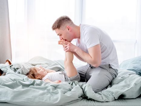 Young father kisses feet of her little daughter lying in the bed. Smiling child with her parent in the morning time. Beautiful moments of dad with his kid in the bedroom together