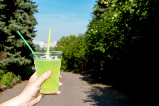 Woman holding plastic green cocktail cup close-up, outdoors in summer in the park.