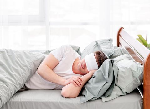 Young man sleeping in the bed under the blanket in stylish interior. Guy lying and napping in the room with sunlight in the morning. Concept of resting time