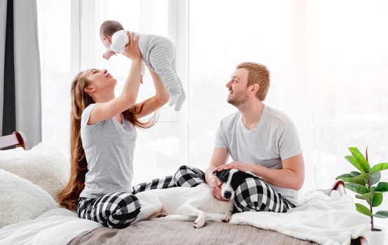 Family with baby boy sitting on the bed with cute dog. Mother holding son in hands and father looking at them. Beautiful parenthood time. Pet with owners