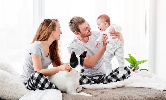 Happy family with baby boy sitting on the bed with cute dog. Mother and father with their son and doggy together in room with daylight. Beautiful parenthood time. Pet with owners