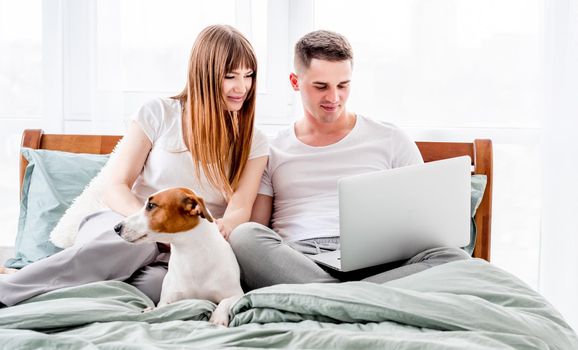 Young couple in the bed with gadgets and cute dog. Beautiful woman and man looking at laptop and girl holding smartphone. Family morning with pet and technologies