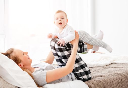 Beautiful young mother holding her adorable little baby boy son on her legs and looking at the child. Pretty girl playing with kid in the bed in sunny room. Happy matherinity moments