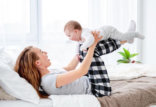 Beautiful young mother holding her adorable little baby boy on her legs and looking at the child. Pretty girl playing with kid in the bed in morning time. Happy family moments