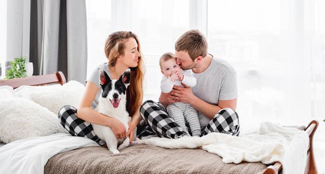 Family with baby boy sitting on the bed with cute dog. Mother hugging doggy and father holding their son and kisses him. Beautiful parenthood time. Pet with owners