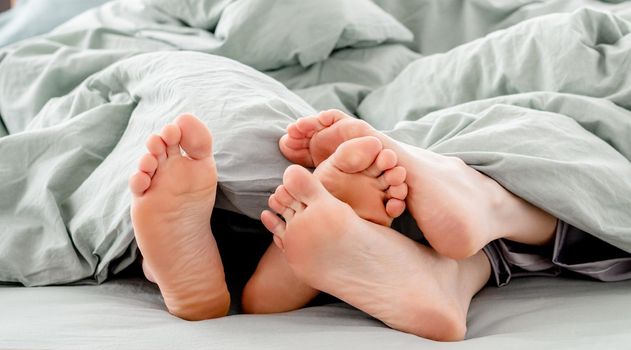 Feet of young couple in the bed under the blanket. Legs of man and woman together touching each other in the bedroom in the morning. Concept of rest, love and happy family relationships
