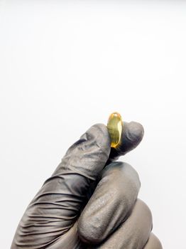 one yellow capsule of fish oil in hand in a black medical glove on a white background. Hand with pills on a white background