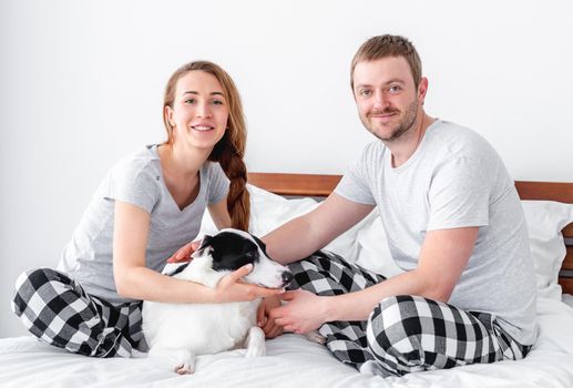 Beautiful caucasian couple sitting in the bed with cute dog. Young man and woman with pet looking at the camera and smiling. Portrait of happy family with doggy