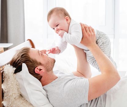 Young father lying in the bed with little son in the sunny morning time and holding adorable kid in his hands. Man with child baby boy playing in the bedroom. Happy parenthood closeup portrait