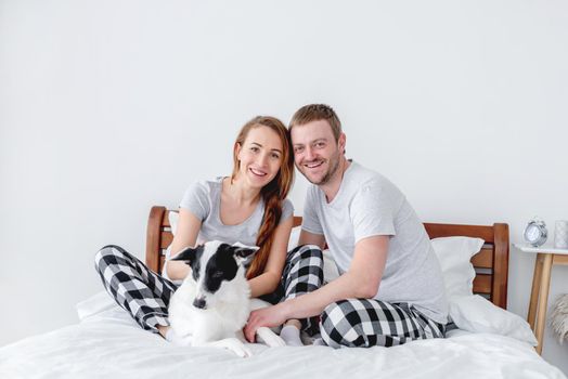 Beautiful caucasian couple sitting in the bed with cute dog. Young man and woman with pet looking at the camera and smiling. Concept of happy family and love