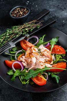 Salad with baked salmon fillet steak, fresh arugula and tomato in a plate. Black background. Top view.