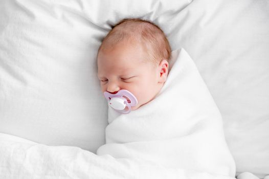 Adorable newborn baby girl swaddled in white blanket sleeping with soother in the bed with white sheets. Cute infant child napping at home