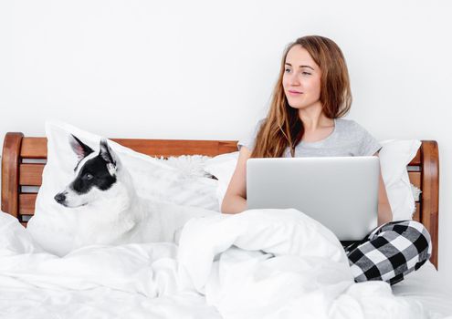 Beautiful girl wearing pajamas sitting in the bed with laptop and cute doggy lying close to her. Young woman at home and pet looking back together in morning time. Concept of freelance