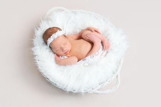 Small child napping naked in a basket, wearing a white flowery hairband