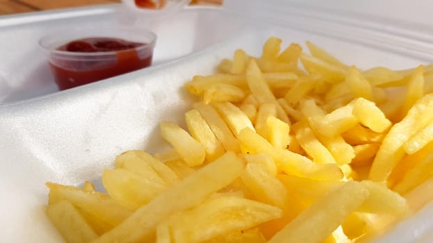A close-up of golden orange yellow deep fries with tomato ketchup served in a foam container. Fast food takeaway. Unwanted fresh food