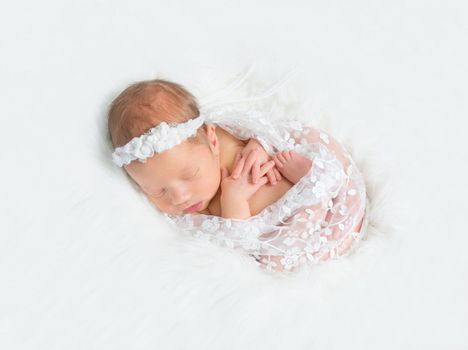 Lovely baby sleeping tightly wrapped with a blanket, in a hairband on white furry surface