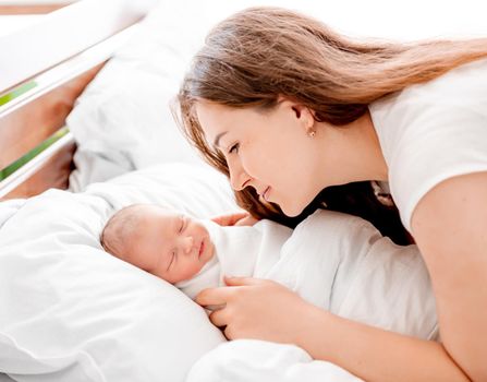 Young mother looking at her sleeping newborn daughter swaddled in white sheets at home. Portrait of girl mom with her infant child napping in the bed