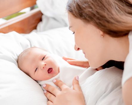 Young mother looking at her awaking newborn daughter swaddled in white sheets at home and lying with mouth open. Portrait of girl mom with her infant child in the bed